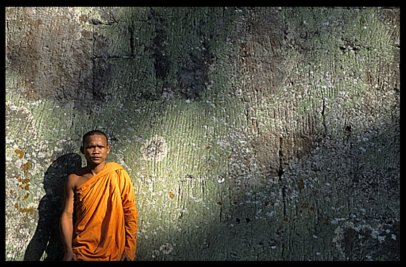 A monk is resting against a weather beaten wall inside Ta Phrom.