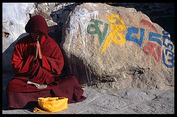 A praying Tibetan monk next to a mani stone carved with sutras in Drepung Monastery.